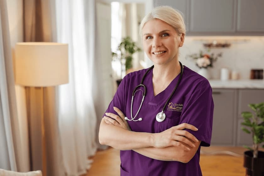 A caregiver in purple scrubs standing with folded arms in a home, smiling.