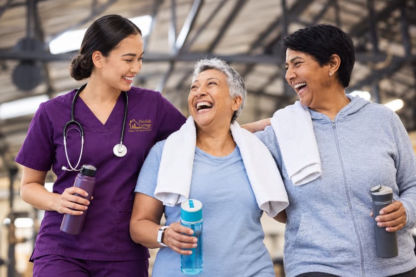 A caregiver in purple scrubs walking and laughing with two older women after exercise.