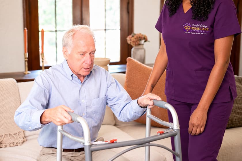 A caregiver supports an older man as he rises from a sofa, both hands on his walker.