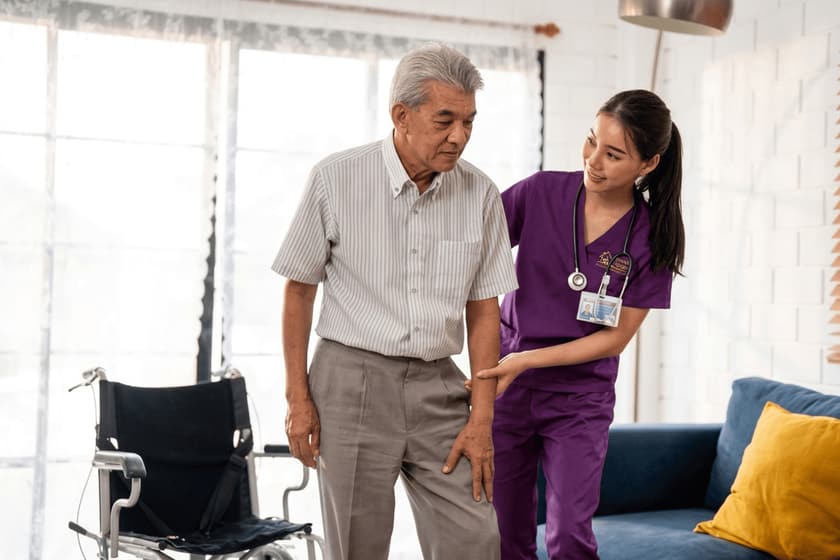 A caregiver walks beside an older man across a living room, a wheelchair resting behind them.