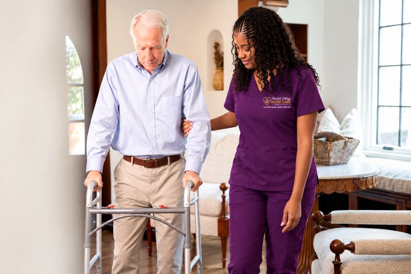 A caregiver walks beside a man using a walker in a bright living room.