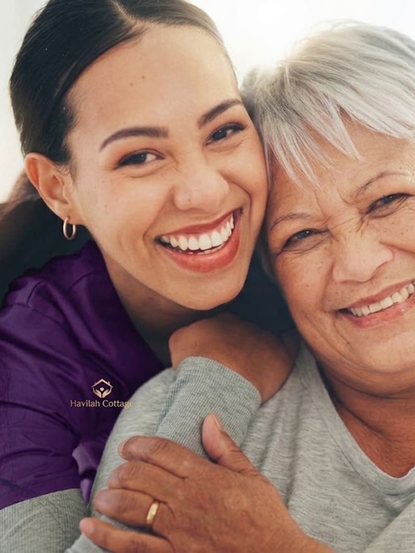 A caregiver and an older woman smiling warmly cheek to cheek.